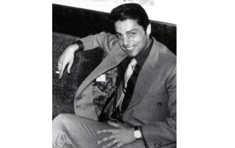 A black-and-white archival photo of a man in a suit and tie reclining with his arm propped up on the back of the chair he sits on. He smiles at the camera. 