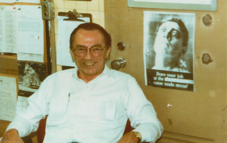 This historical photo shows Robert Kirkpatrick in a white collared shirt in his office at the World Trade Center. Papers and images are tacked onto the wall behind him.  