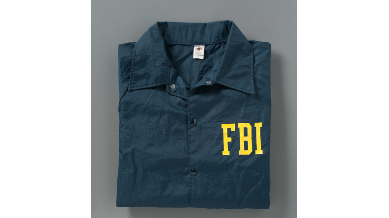 A navy blue windbreaker with "FBI" printed in yellow lettering on the left pocket rests neatly folded on a gray background. 