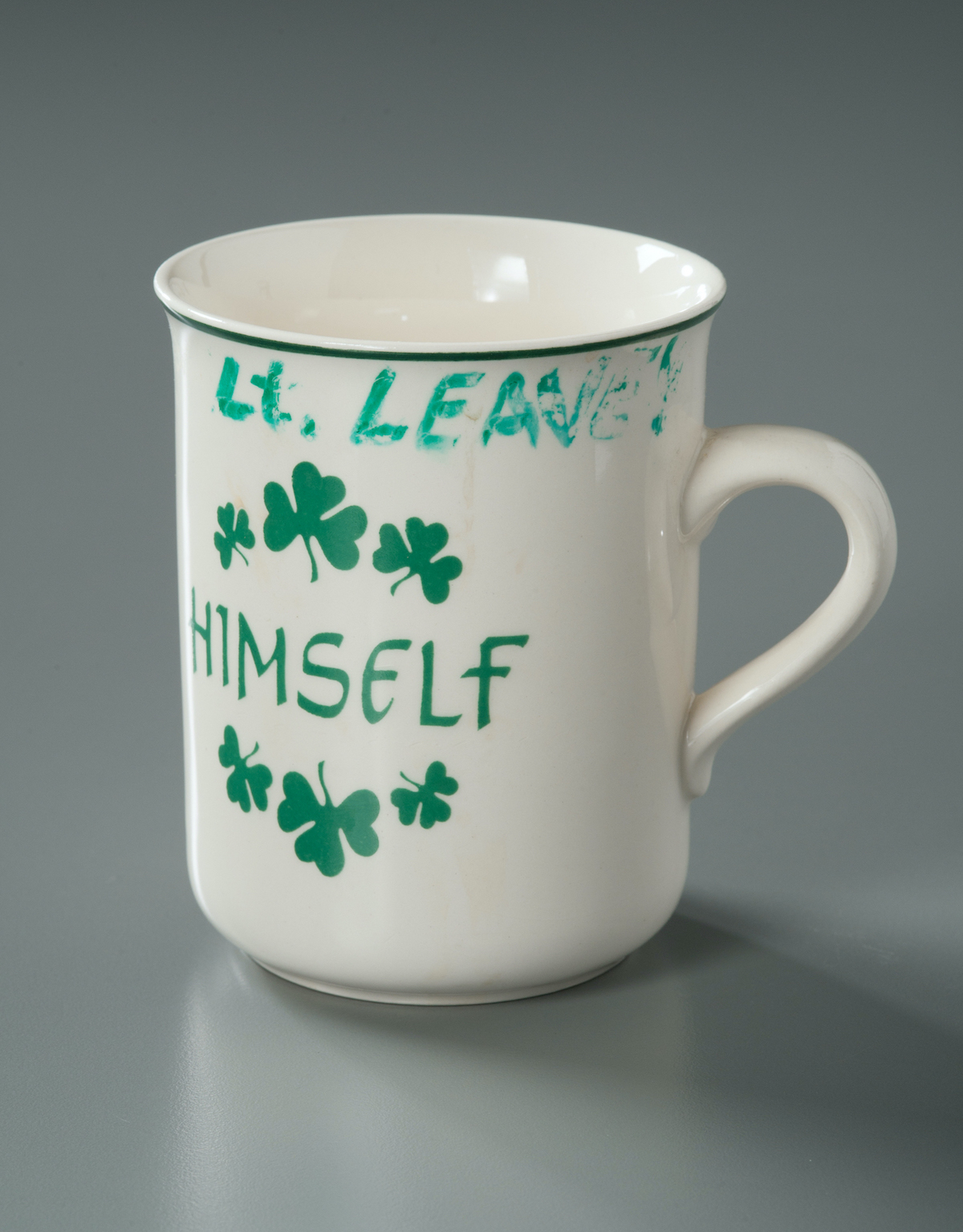 A mug belonging to FDNY Lieutenant Joseph Leavey is displayed on a gray surface at the museum. The white mug is adorned with green shamrocks. The word "himself" is printed in green on the mug. Leavey's name has been written in green marker on the lip of the cup. 