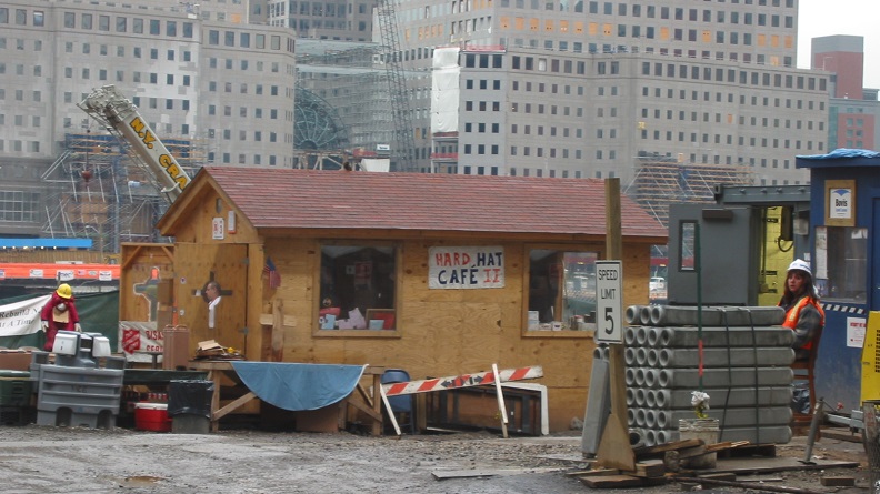 This photo shows a plywood shack in the middle of Ground Zero with a sign reading "Hard Hat Cafe" affixed to the outside. 