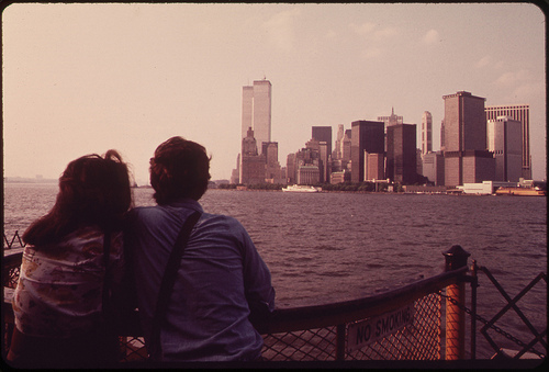 A man and a woman standing on the back of a boat on a calm water, facing towards the Lower Manhattan skyline. The Twin Towers is visible alongside other skyscrapers. 