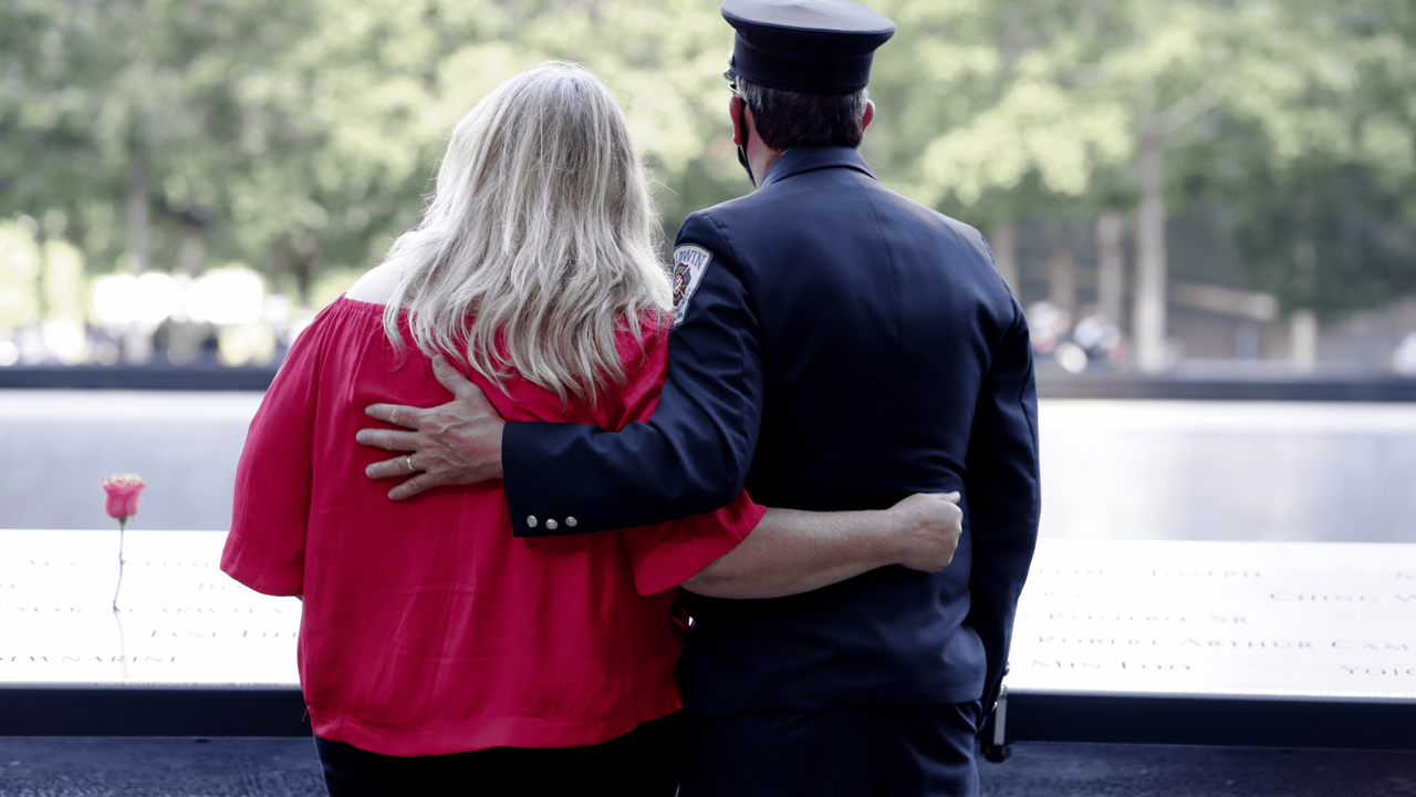 A woman and a man wearing firefighter dress blues gaze over the names panel and into the pool of the 9/11 Memorial Museum on the 19th Anniversary of the attack.  The man has his arm draped over the woman's shoulder.  She has her arm around his waist. 