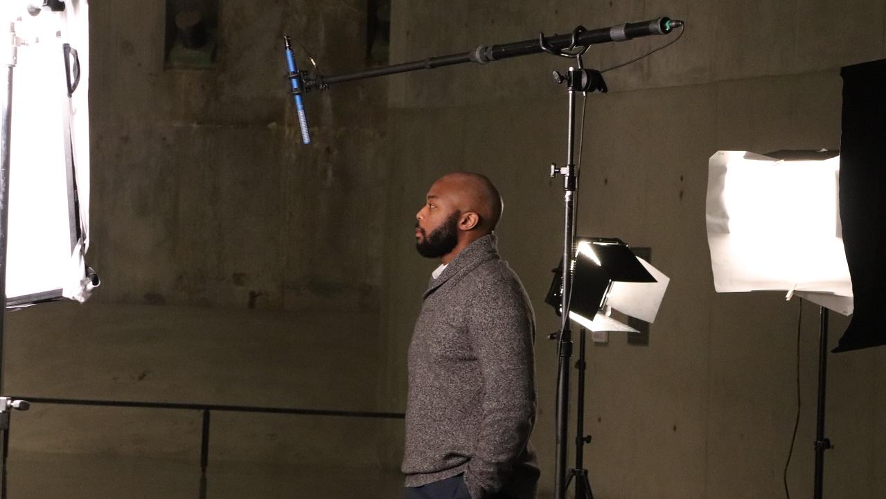 In this photograph a man stands in profile before a  boom mic and set lights during the Anniversary in the schools webinar filming 