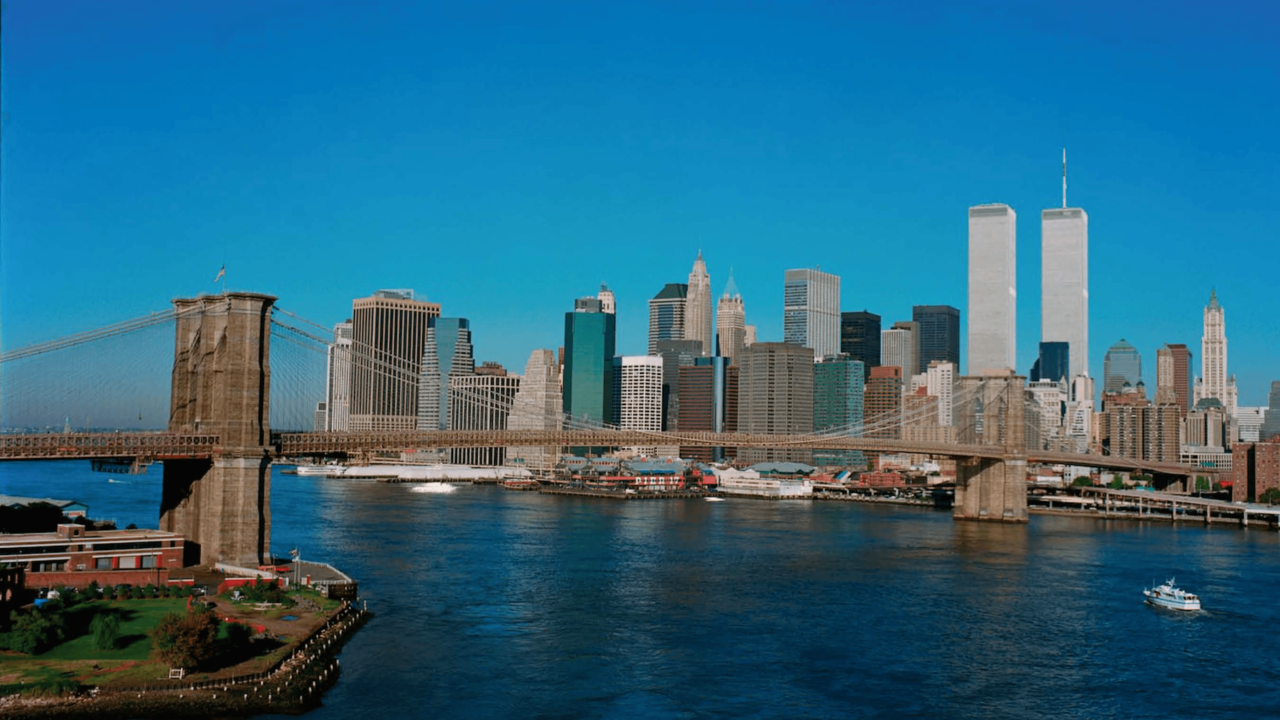 View of lower Manhattan's building behind a bridge spanning a calm river. The Twin Towers stand tall above other buildings, reaching up to a clear blue sky.  