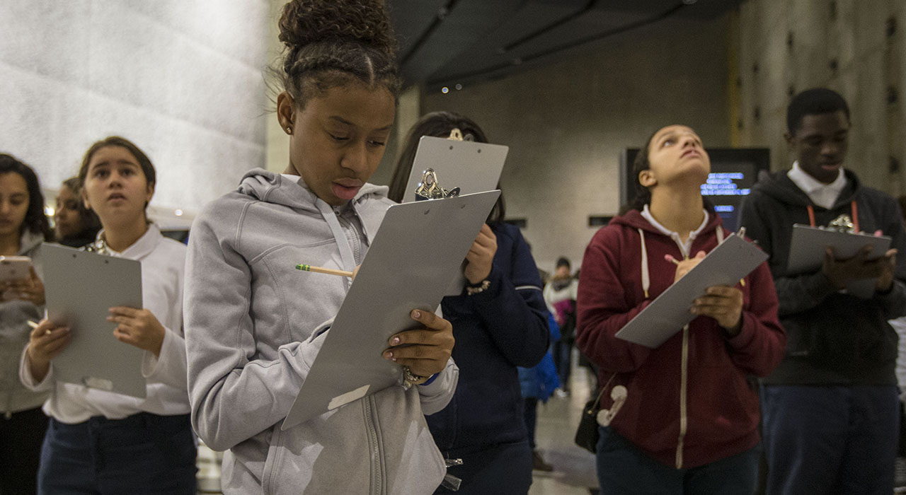 Group of middle school students in different skin tones write on clipboards inside the museum, with aluminum cladding visible in the background. 