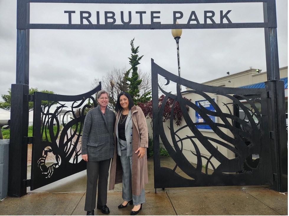Elizabeth Hillman and Joann Ariola pose in front of a decorative black metal gate that says Tribute Park 