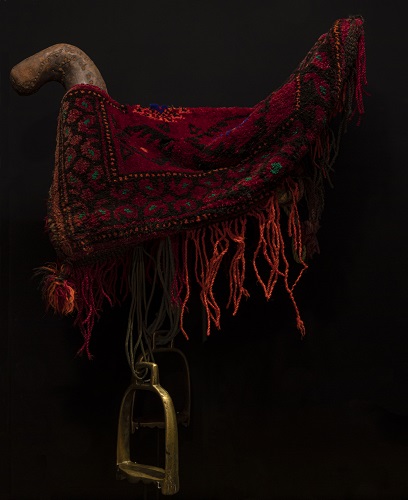 Saddle with metal stirrup pads draped in a short red rug decorated with patterns and fringes.