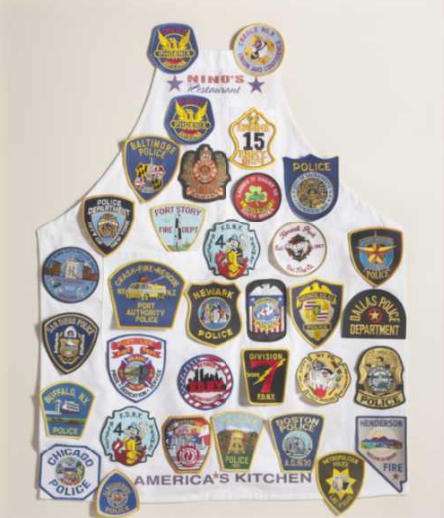A white apron covered in 34 uniform patches representing a variety of rescue and recovery agencies at Ground Zero. "Nino's Restaurant" is printed at the top of the aprong and "America's Kitchen" at the bottom.