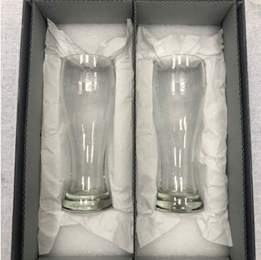 Two glass pint glasses sit side-by-side in a gray box resting on a bed of white tissue paper.