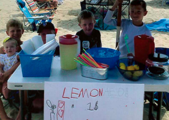 Small children selling lemonade at white folding table covered in colorful straws, cups, and pitchers
