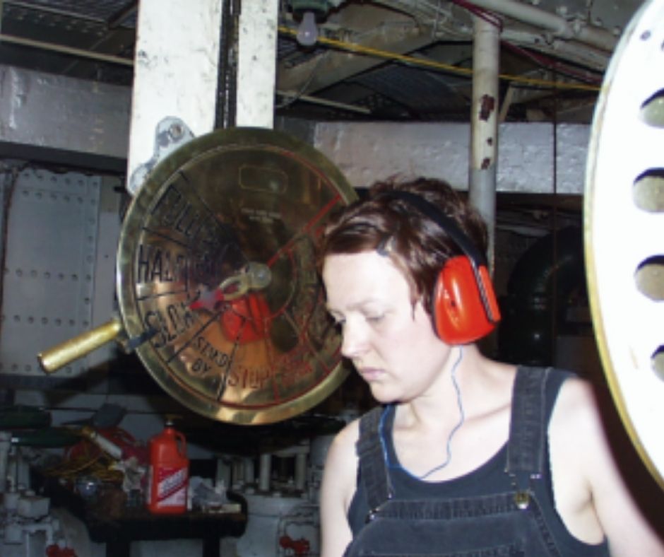 A woman with cropped hair wearing a black tank top and red earphones while working in a ship's engine room