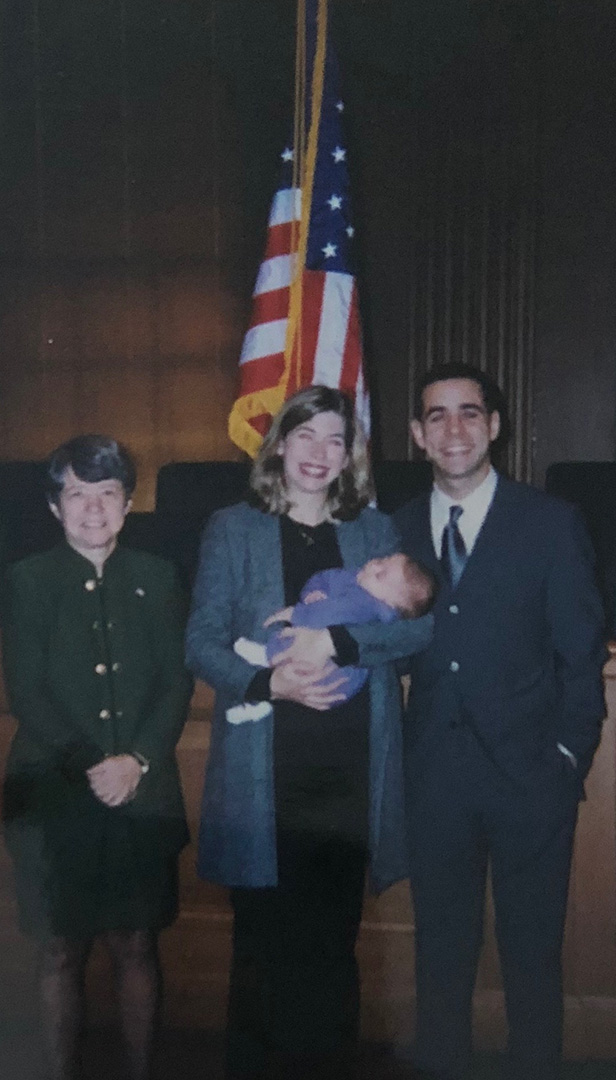Three smiling adults standing in front of an American flag. The woman in the center cradles a baby.