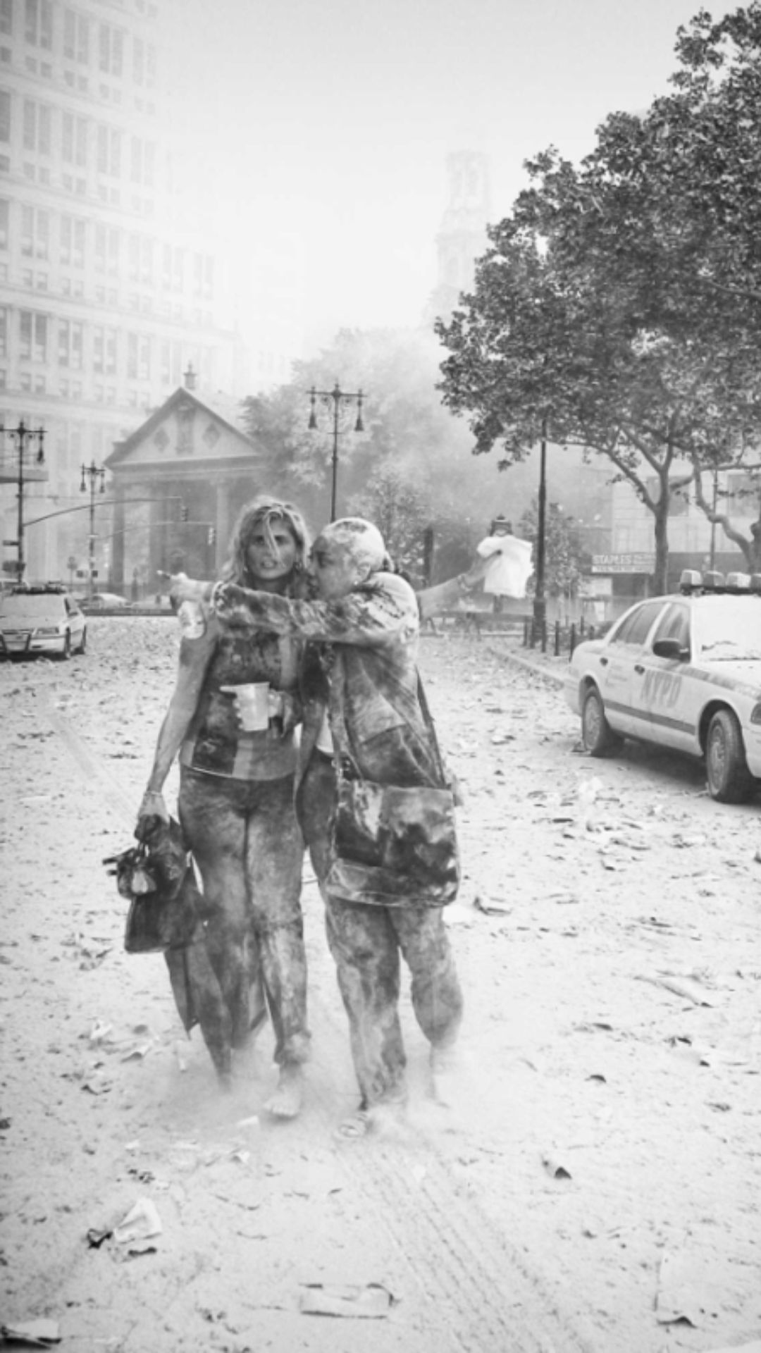 A pair of women in the foreground, covered in dust, as additional unsettled dust fills the air and the street is covered in debris.