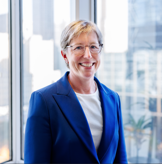 Beth Hillman, our President and CEO, smiles in front of two large windows. She is wearing a bright blue blazer with a white top underneath, and glasses.