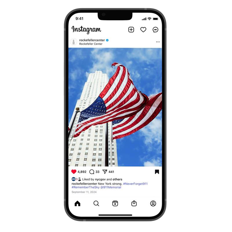 Two American flags blow in the breeze with 30 Rockefeller Plaza and a bright blue sky, peppered with white clouds, in the background