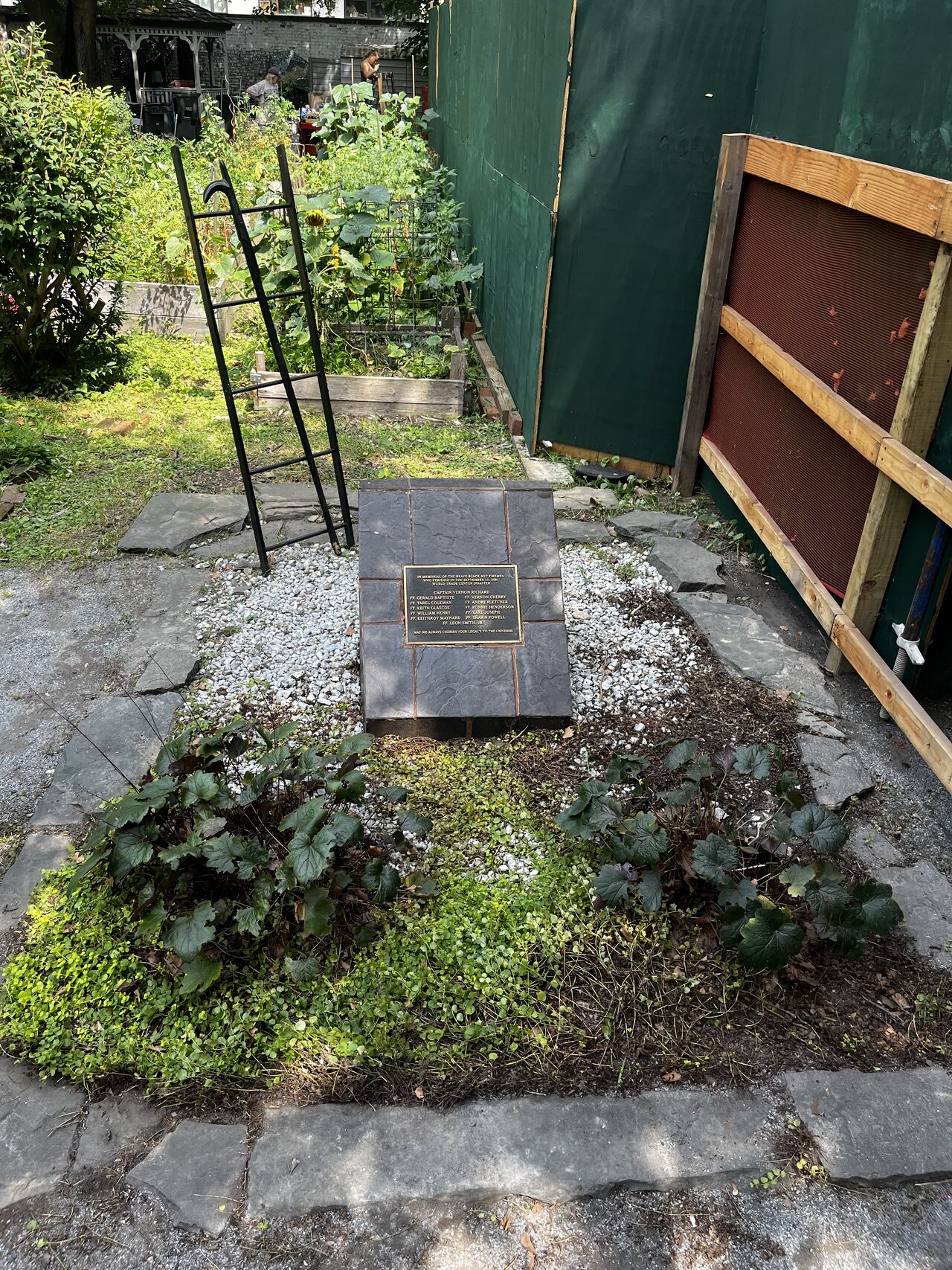 A bronze and black engraved plaque atop a short slate pillar in a small garden. A metal ladder extends from behind the pillar.