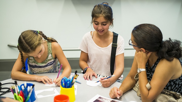 Two girls are engaged in an art activity in a classroom setting while a woman sharing their table looks on attentively.