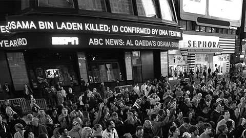 Back view photograph of a standing crowd in the middle of the street. News ticker above announces Osama Bin Laden Killed.