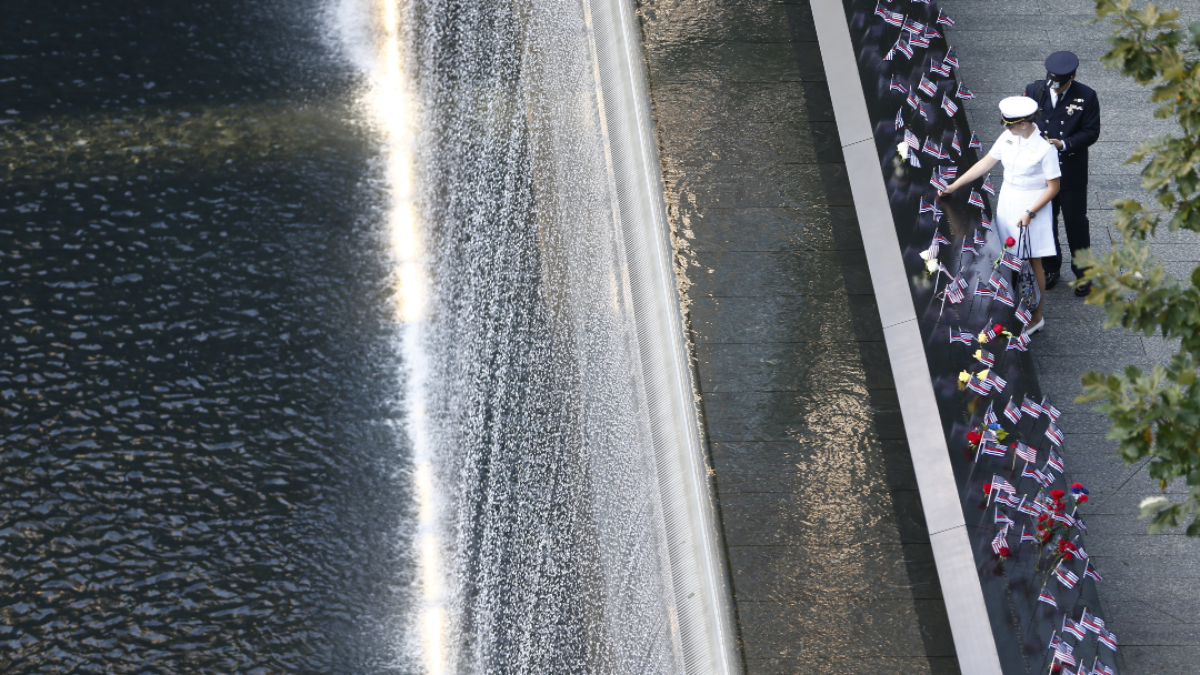 The overview shot of the 9/11 Memorial depicts the water cascading down the Memorial.  In the upper right corner, stand two people wearing military uniforms.  A man in dress whites adds an American flag to a name in a parapet.  The parapet is filled with red, white and blue.  