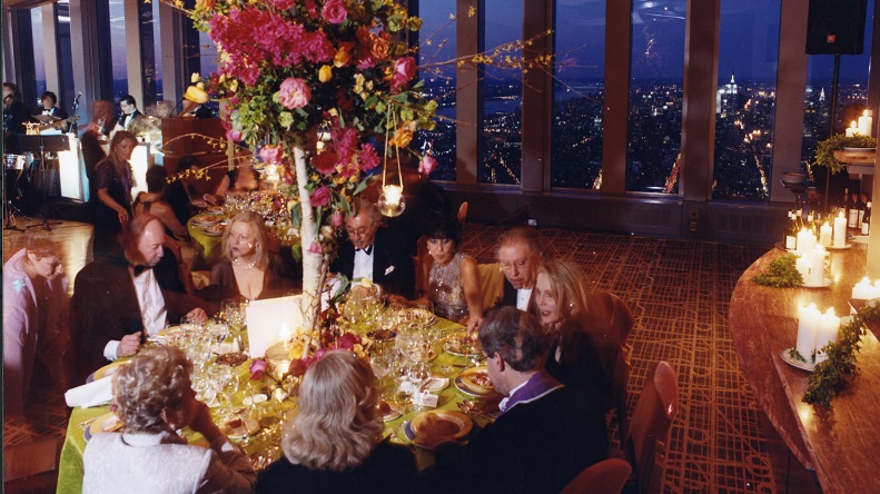 Photograph with high-angle view of people in formal wear sitting around a table with high floral centerpiece. Rows of full length windows in the background show glimpse of nighttime aerial city view.