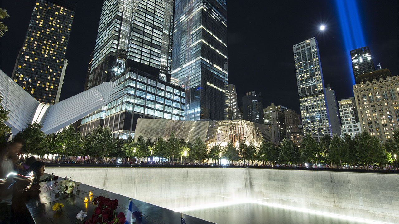 Visitors surround the brightly lit north reflecting pool at night. The Museum pavilion and nearby skyscrapers are also alight. A radiant moon and the blue glow of the Tribute in Light shine above the buildings.