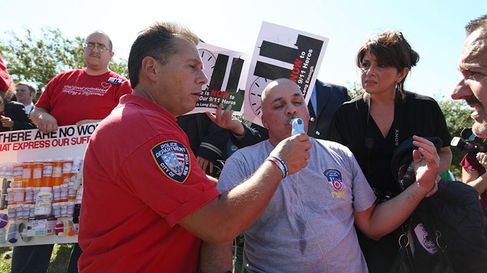 Paramedic administrating inhaler to a man during a rally. Multiple signs behind them show images of prescription vials and black silhouette of the Twin Towers.