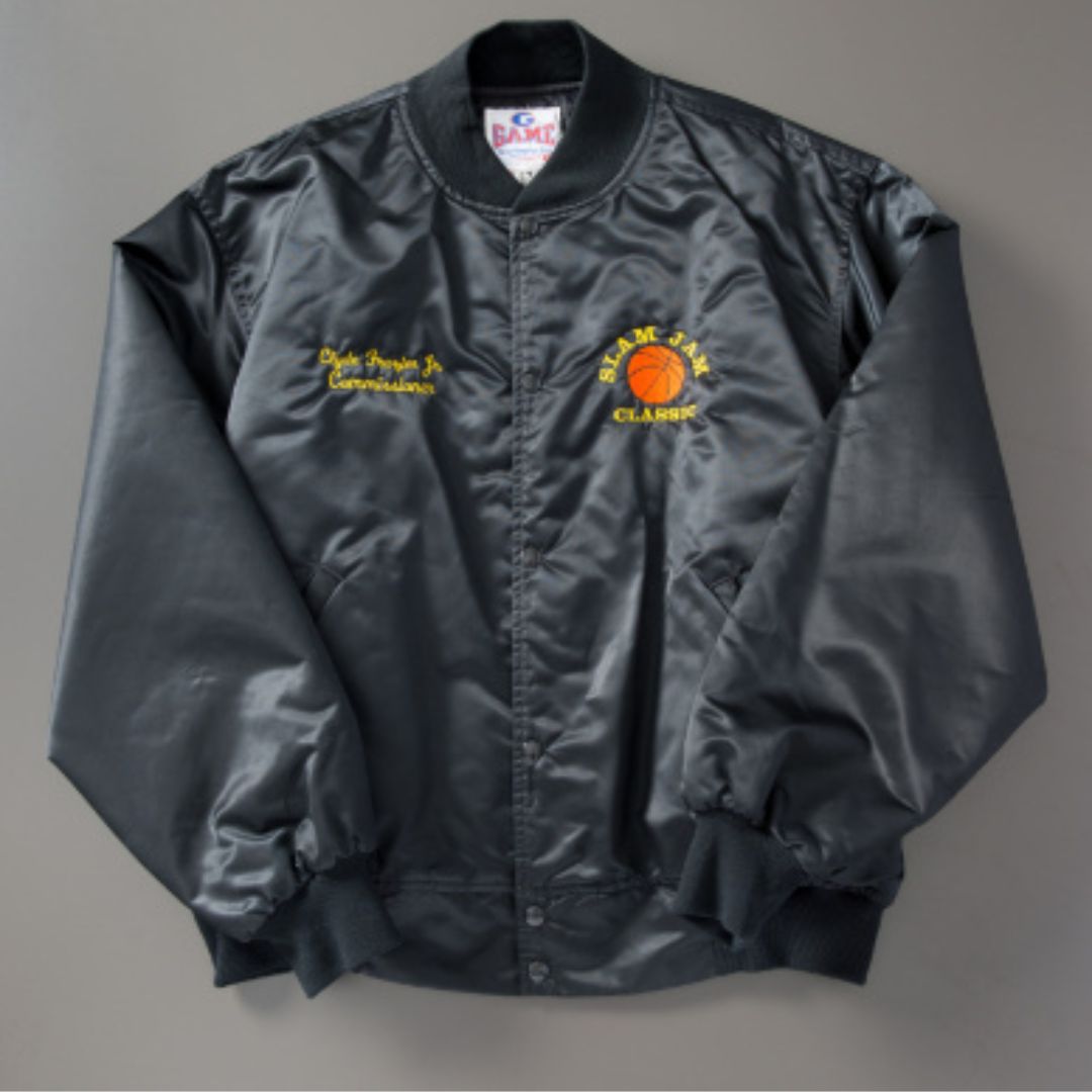 A black satiny basketball jacket against a gray background. The jacket has bright yellow stitching on it that reads (left) Clyde Frazier Jr., Commissioner, and (right) Slam Jam Classic, which wraps around a basketball.