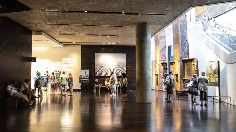 Visitors explore the lobby of the Museum. The gift shop is off to the left, twin steel beams called the Tridents are to the right, and a large image of one of the Twin Towers is at the center. 