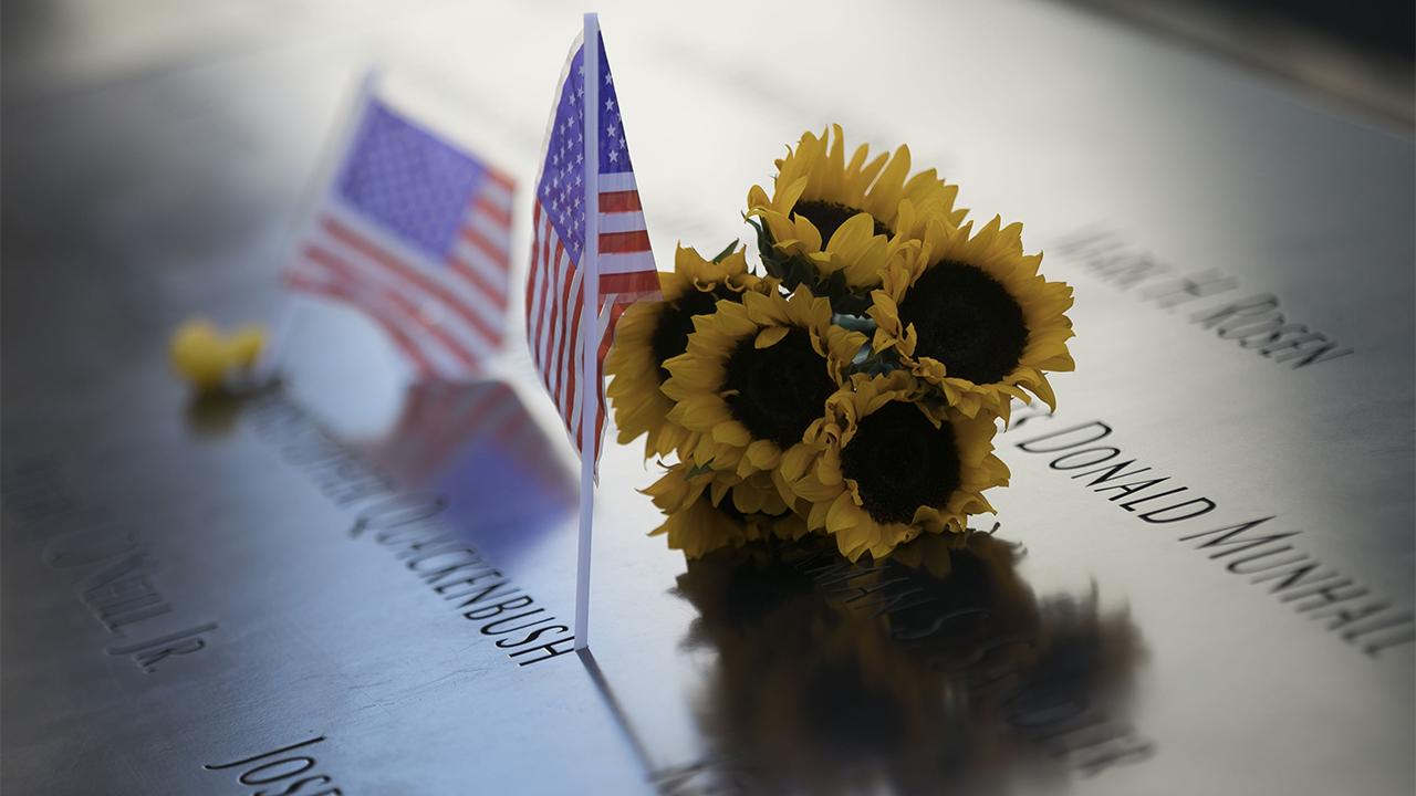 A bunch of sunflowers rest on the 9/11 Memorial, which is cast in a shimmery yellow and blue light. Two American flags rest alongside it.