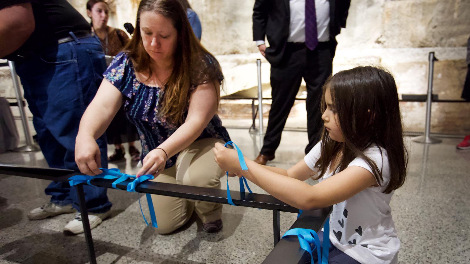 A woman and a young girl kneel down as they place blue ribbons on a black railing at the foot of the Last Column. Several other people stand behind them.