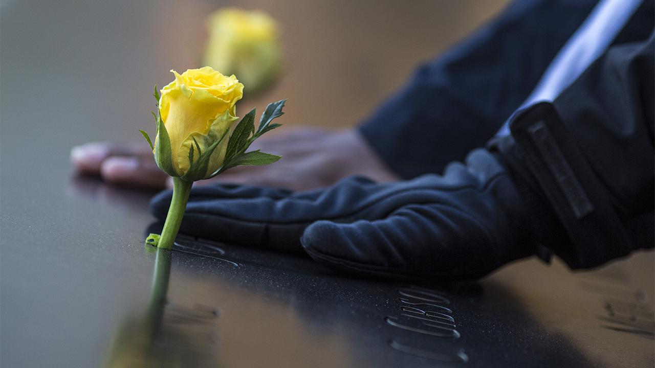 A person’s gloved hand touches a name adorned with a yellow rose on a bronze parapet at the Memorial. The hand of a second person is between the first hand and a second yellow rose.