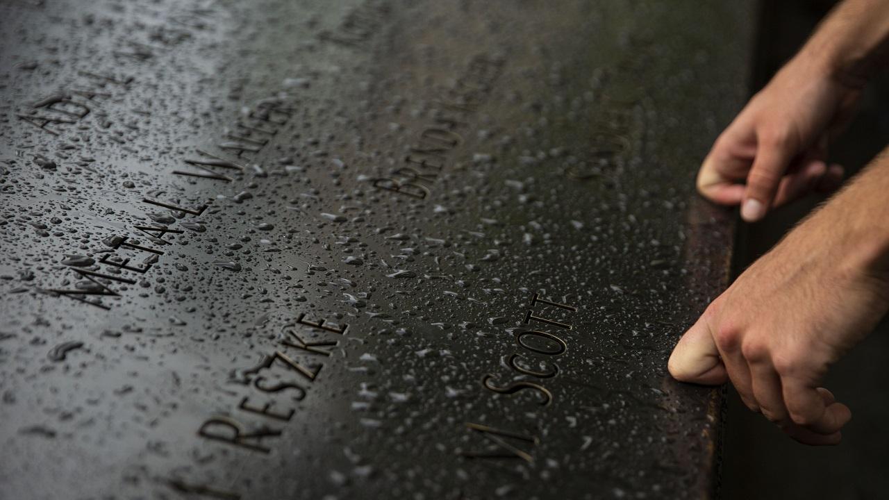 A person’s hands are placed on a bronze parapet covered in raindrops. The knuckles of the index fingers are bent against the parapet next to two victims’ names.