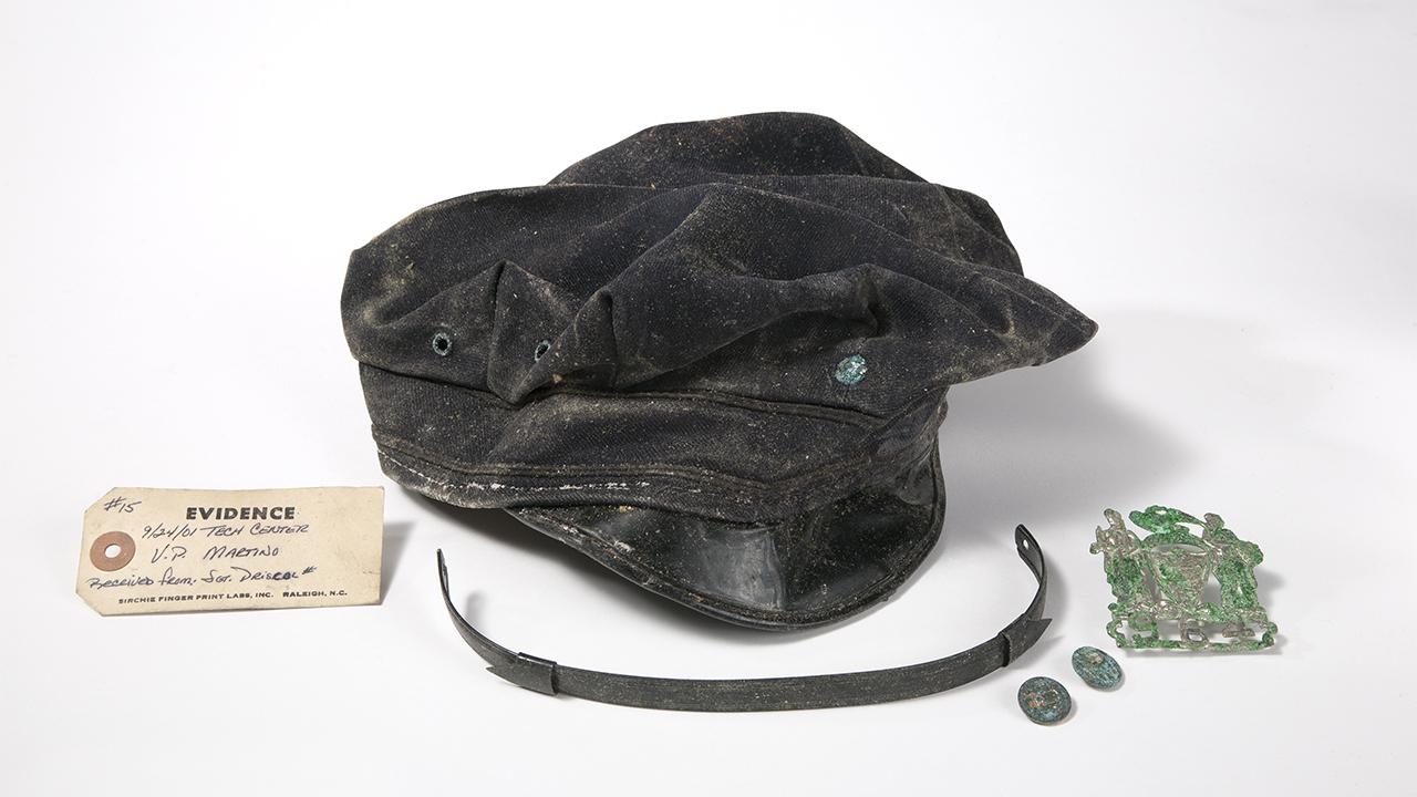 Several damaged items belonging to Port Authority Police Officer Sharon Miller are displayed on a white backdrop with a tag marking them as evidence. The items include a misshapen police cap and a damaged copper badge.