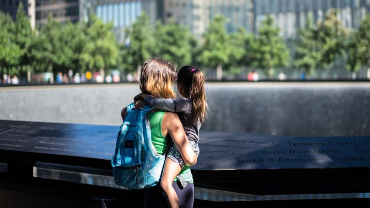A woman with a backpack holds a young girl with a ponytail as they both look at one of the Memorial’s reflecting pools on a sunny day. Green oak trees and buildings are seen farther afield.