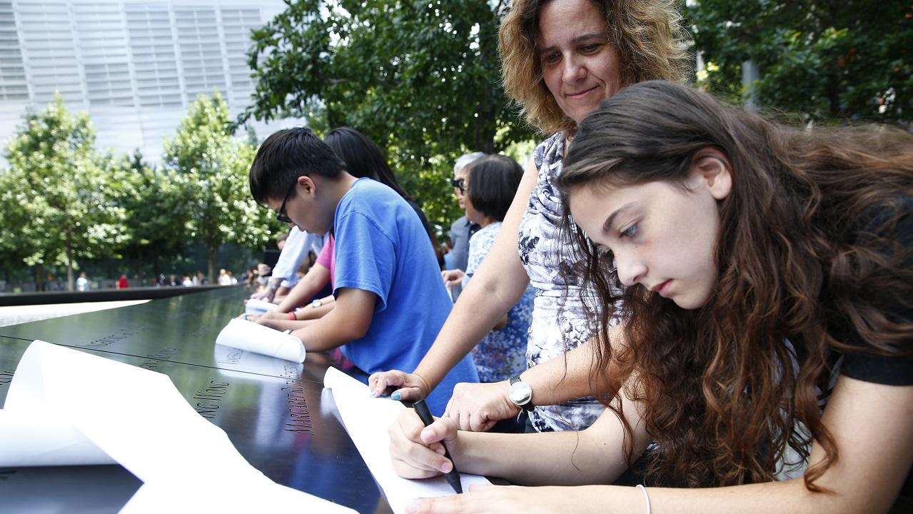 A teenage girl and a woman create a name impression at the Memorial by rubbing charcoal over a sheet of paper placed on an inscription. Other families beside them do the same along a bronze parapet inscribed with victims’ names.