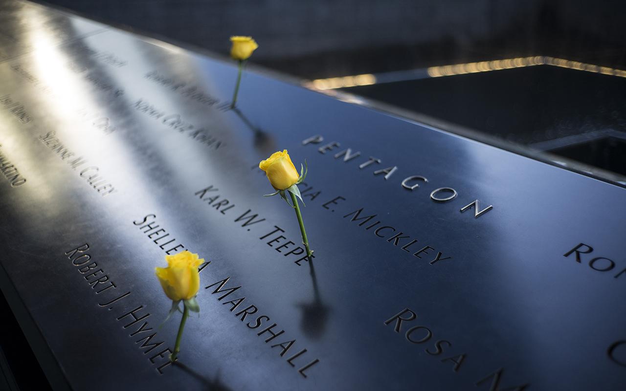 Three yellow roses stand at the names of victims etched on a sunlit Memorial. The names are near the inscription indicating Pentagon victims. The water of a reflecting pool can be seen further afield.