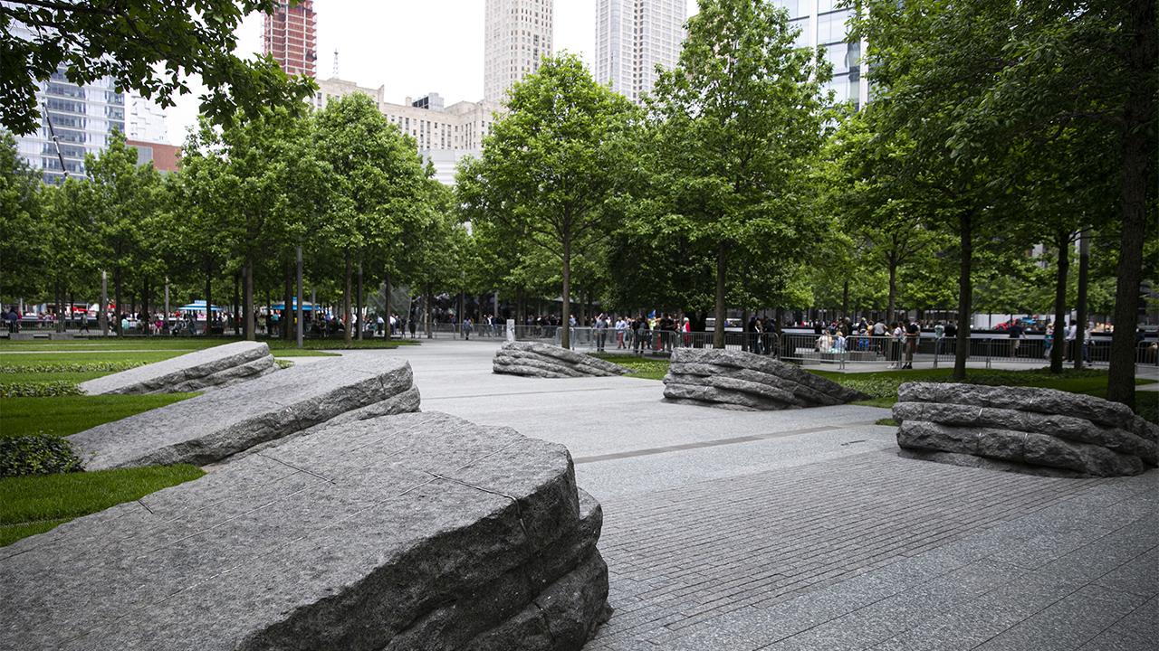 Six stone monoliths border the pathway of the 9/11 Memorial Glade. There are three monoliths on each side of the path. Trees with bright green leaves stand to the left and right and also off in the distance.