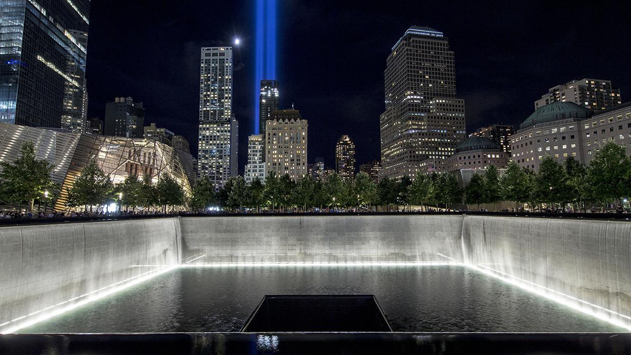 Water cascades down the illuminated walls of the North Tower reflecting pool on a warm night. The water pours down a square hole at the center of the pool. In the distance, a moon hangs over the city and the Tribute in Light shines above the buildings.