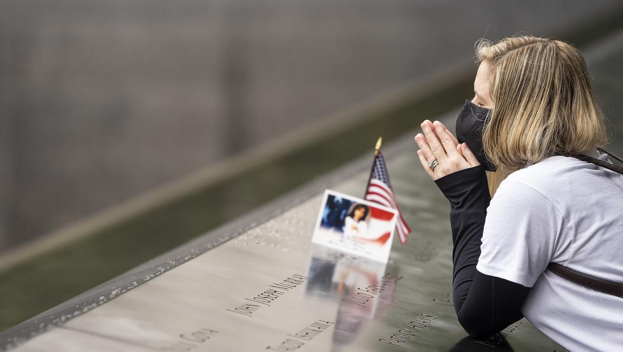 A women holds her hands, clasped in prayer, near her face as she leans on the 9/11 Memorial.  A name on the Memorial is adorned with a photograph and American flag as this visitor closes her eyes in remembrance and prayer.