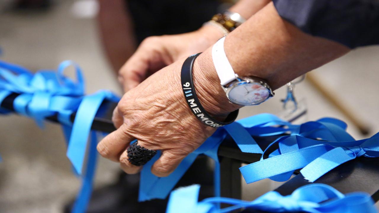 In this close-up image, a pair hands, adorned with a 9/11 Memorial Museum bracelet, are seen tying a blue ribbon to the Last Column in Foundation Hall of the 9/11 Memorial Museum.