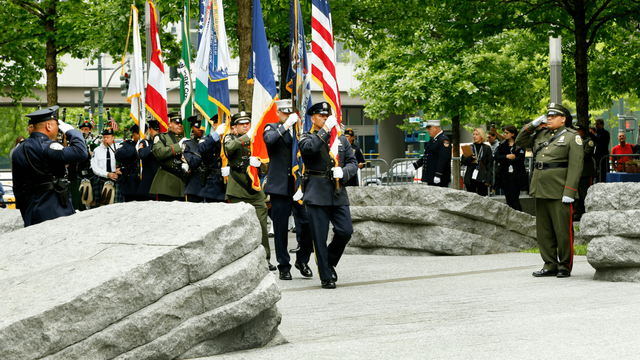 A color guard of more than 12 people enter the Memorial Glade carrying the American and New York State flags among others enters the Memorial Glade on a sunny day as two men in uniform salute.  