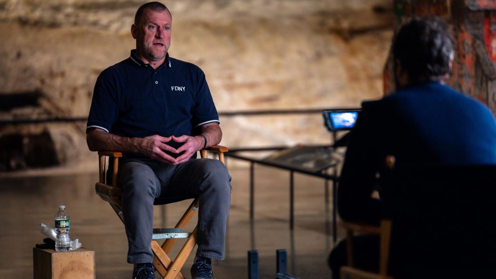 A person wearing jeans and a blue FDNY polo shirt sits in a director's chair in front of the Slurry Wall. Another person, slightly out of focus, is interviewing him.