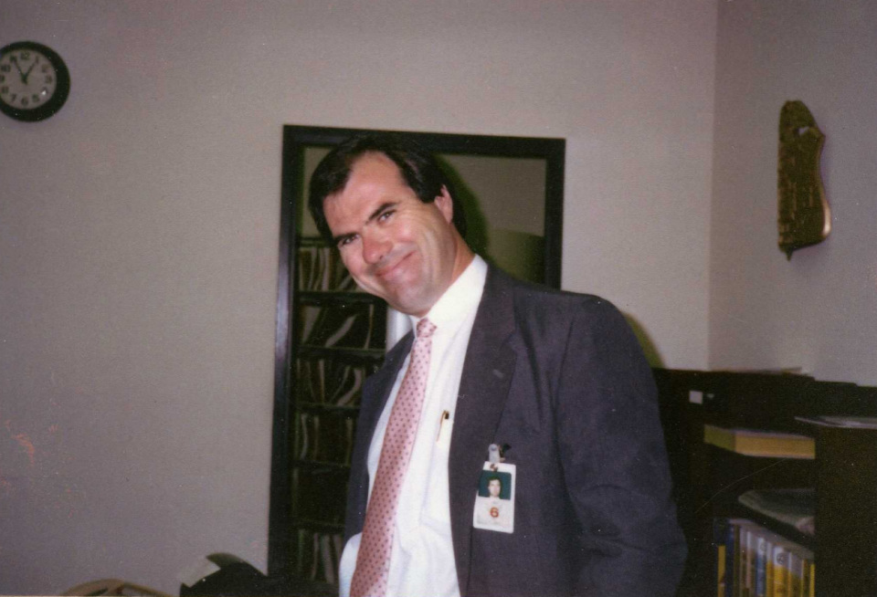 A man wearing a suit and tie and a professional badge grins with his head tilted to the side in an office.