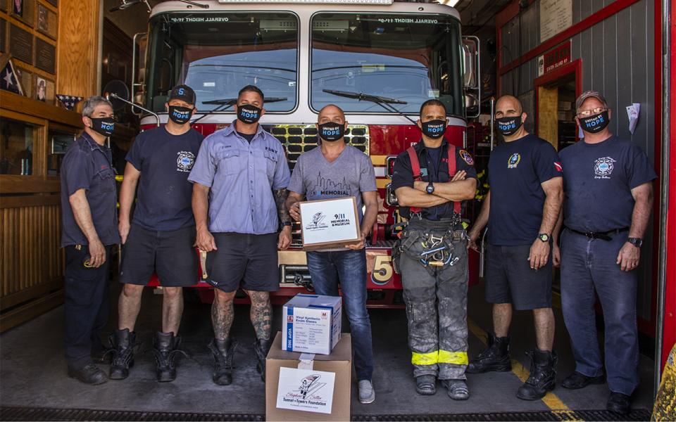 Six firefighter stand in front of a firetruck at a firehouse on Staten Island.   In the center of the firemen, an employee of the 9/11 Memorial Museum stands holding a box of masks that were delivered to the fire house. 