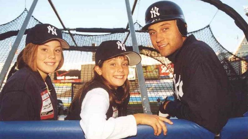 Two young women sit with New York Yankee Derek Jeter on a bench, where they are turned around to pose for the camera behind them.