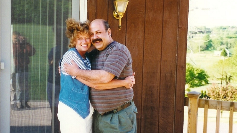 A man and a woman stand on a deck, hugging each other with big smiles.