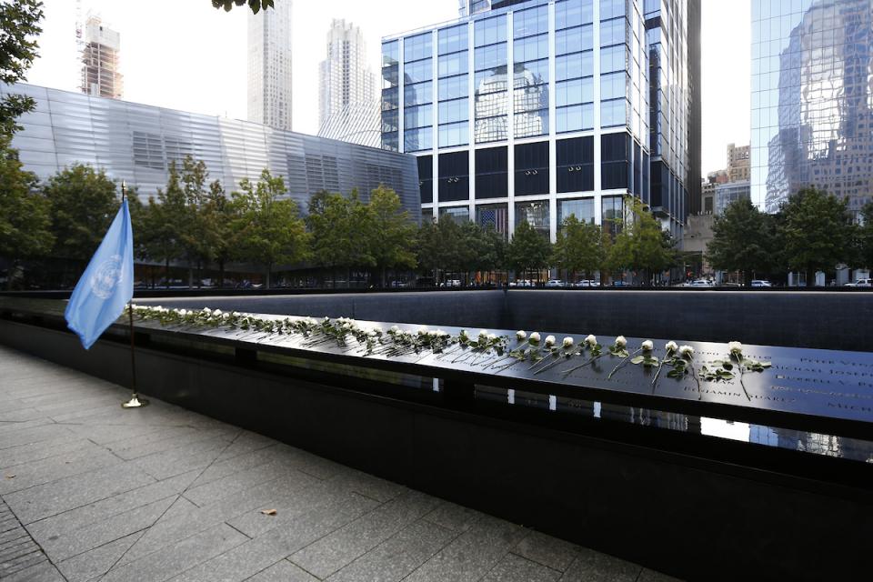 The United Nations flag stands to the left of the Memorial, which is covered in white roses