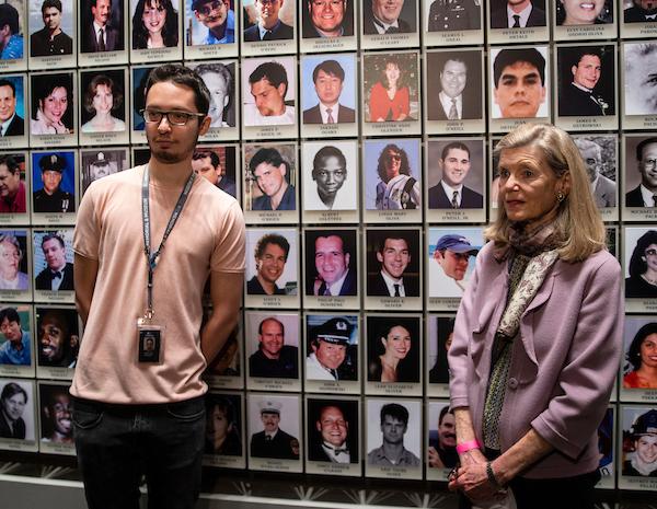 A man in a pink t-shirt (left) and a woman in a lavender sweater stand in front of the Wall of Faces, which features photographs of the victims of the attacks